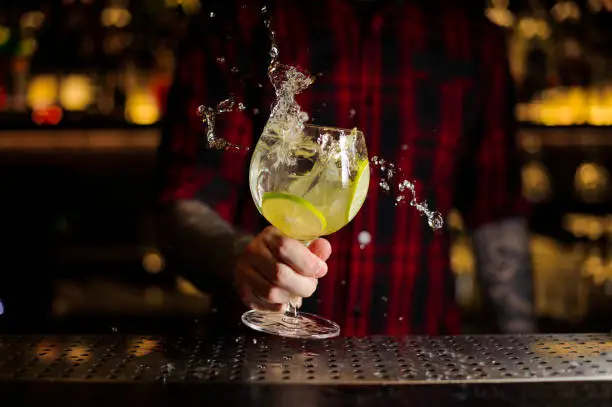 Bartender making splash of a Gin Tonic cocktail with lime slices on the steel bar counter on the blurred background
