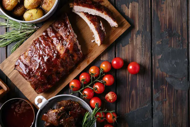 Home made pork and beef ribs, glazed with barbecue sauce served with baked rosemary potatoes, herbs and tomatoes, decorated with napkins over a rustic wooden background. Top View. Copy Space