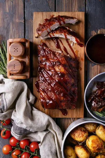 Home made pork and beef ribs, glazed with barbecue sauce served with baked rosemary potatoes, herbs and tomatoes, decorated with napkins over a rustic wooden background. Top View. Copy Space