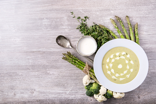 Home made asparagus soup served with cream and thyme, decorated with fresh asparagus, cauliflower and broccoli on grey wooden background. Top View. Copy space