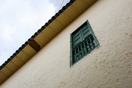 Low angle view of a white rough wall with green wood window and orange shingles roof of a house in Cusco, Peru, with blue sky with clouds on the background.