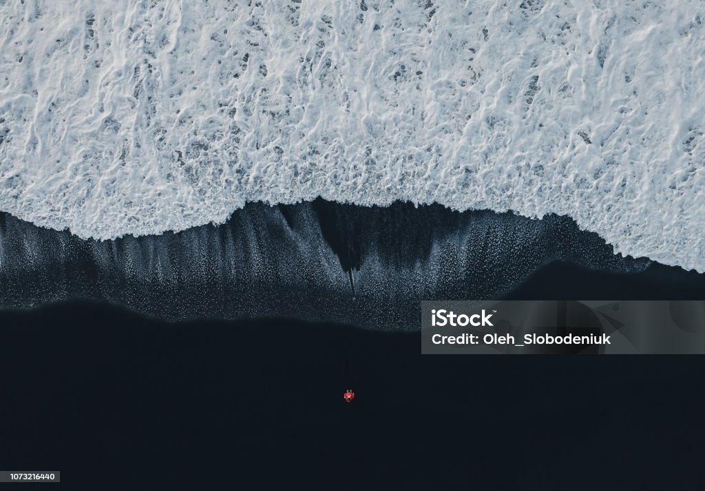 Aerial view of woman on black sand beach in Iceland Aerial view of woman  in red coat standing on black sand beach in Iceland and looking at the sea Wave - Water Stock Photo