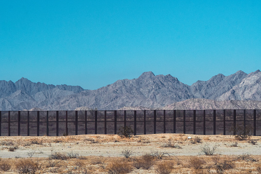The image shows the imposing US-Mexico border wall stretching across the rugged and arid terrain of the border region. The wall, which is made of concrete and steel, rises up to 30 feet in height. It snakes through the landscape, dividing communities and ecosystems alike. The image also captures the stark contrast between the two sides of the border, with the wall standing as a symbol of the ongoing debate over immigration policy in the United States. The image highlights the complexity and controversy of the US-Mexico border region, where issues of immigration, national security, and human rights intersect. Despite its divisive nature, the wall remains a powerful symbol of the challenges facing both the United States and Mexico in the 21st century.