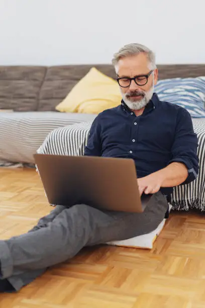 Man relaxing on the floor in his living room at home working on a laptop computer balanced on his lap