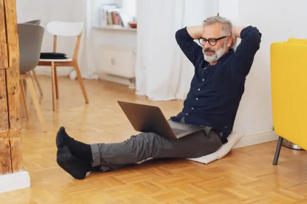 Man relaxing in his socks at home with a laptop sitting on the wooden floor in a doorway in his apartment