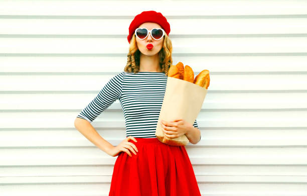 ritratto bella giovane donna che soffia labbra facendo bacio d'aria indossando berretto rosso tenendo in mano sacchetto di carta con lunga baguette di pane bianco su sfondo bianco parete - baguette foto e immagini stock