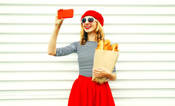 白い壁の背景に長い白パンのバゲットと紙袋を持ってスマート フォンで selfie 写真を撮る陽気な笑顔の若い女性 - フランス文化 写真 ストックフォトと画像