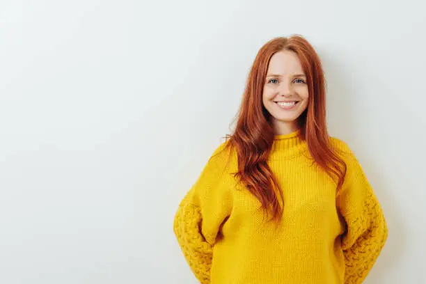 Happy smiling young redhead woman posing in a colorful yellow top against a white wall with copy space