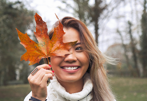 Asian woman holding autumn leaf on her face and smiling in woods