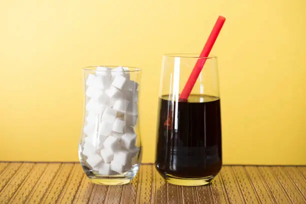 Two drink glasses in front of yellow background.  The first glass is filled with sugar cubes and the second glass is filled with cola and has a red pipette in it.