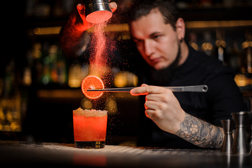 Tattooed professional bartender adding spices powder into a cocktail glass filled with a fresh strong alcoholic cocktail with slice of lemon