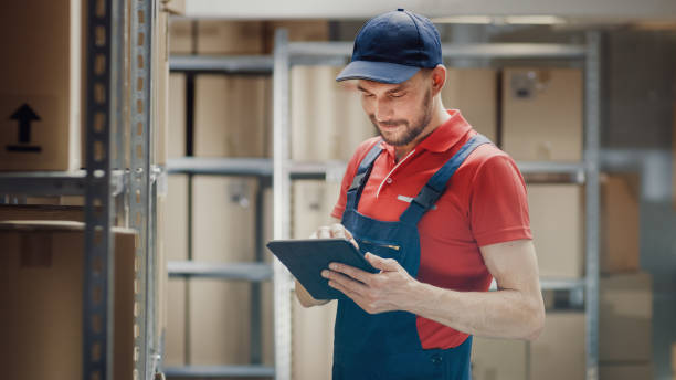 istock Handsome Warehouse Worker Uses Digital Tablet For Checking Stock, On the Shelves Standing Cardboard Boxes. 1073311602