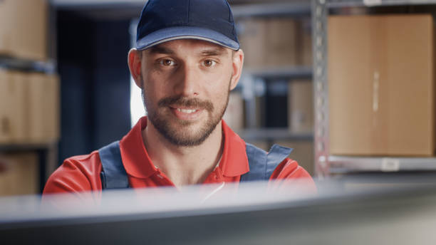 istock Portrait of Uniformed Worker Using Personal Computer while Sitting at His Desk in the Warehouse. 1073311932