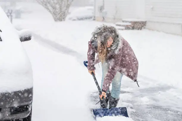 Young woman, female in winter coat cleaning, shoveling driveway, street from snow in heavy snowing snowstorm, holding shovel, residential houses, by cars parked on road