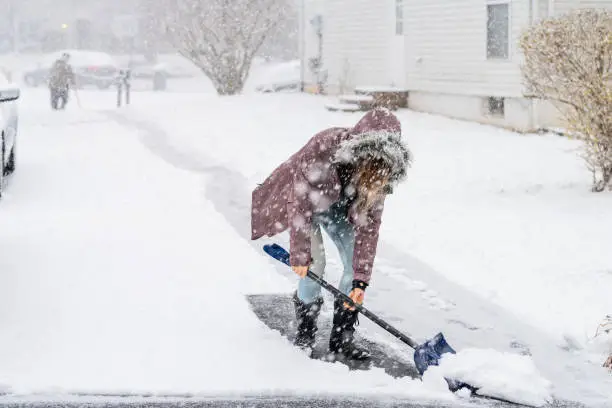 Young woman, female in winter coat cleaning, shoveling driveway, street from snow in heavy snowing snowstorm, holding shovel, residential houses, cars parked on road