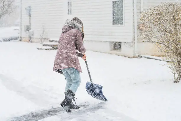 Back, side of young woman, female in winter coat cleaning, shoveling driveway, throwing snow on street in heavy snowing snowstorm, holding shovel, residential houses