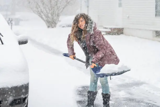 Young woman, female in winter coat cleaning, shoveling driveway, street in heavy snowing snowstorm, throwing, holding shovel with heap, pile of snow, residential houses, by cars parked on road