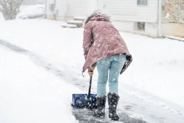 Back of young woman, female in winter coat cleaning, shoveling driveway, street from snow in heavy snowing snowstorm, holding shovel, residential houses