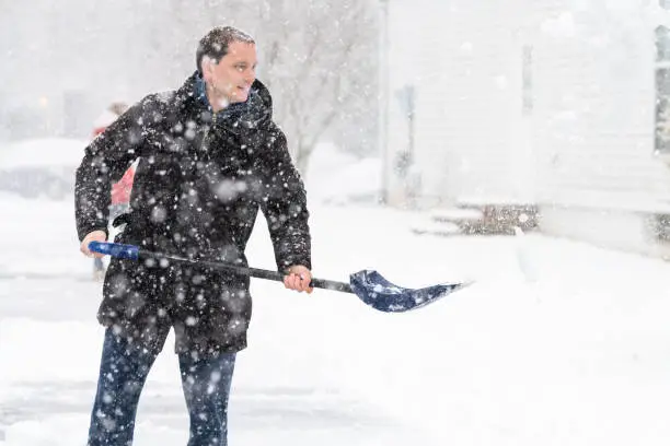 Happy, smiling young man, male in winter coat cleaning, throwing, shoveling driveway, street from covered falling, snow in heavy snowing snowstorm with shovel