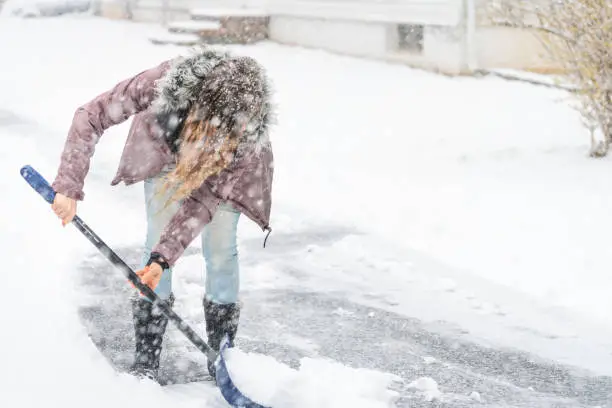 Young woman, female in winter coat cleaning, shoveling driveway, street from falling snow in heavy snowing snowstorm, holding shovel, residential houses