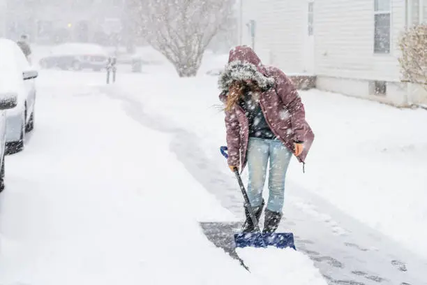 Young woman, female in winter coat cleaning, shoveling driveway, street from snow in heavy snowing snowstorm with shovel, residential houses, cars parked on road