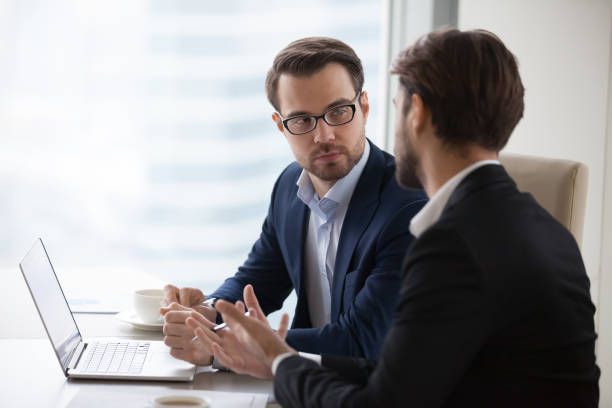 Two young businessmen discuss or plan project in office Two serious caucasian men in suits discussing or planning business issues in the office. Colleagues or client and consultant are sitting at the table next to each other and talking. serious business stock pictures, royalty-free photos & images