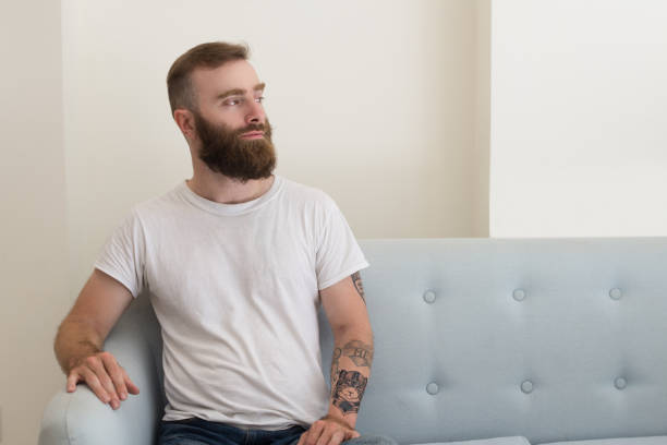 istock Pensive handsome bearded man in white tshirt sitting on sofa 1073442978