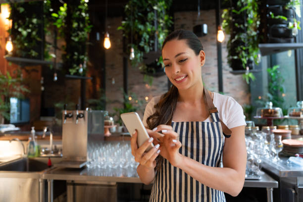 cameriera allegra latinoamericana che si prende una pausa chiacchierando su smartphone sorridendo mentre si trova dietro il bancone del bar - smiling women glasses assistance foto e immagini stock