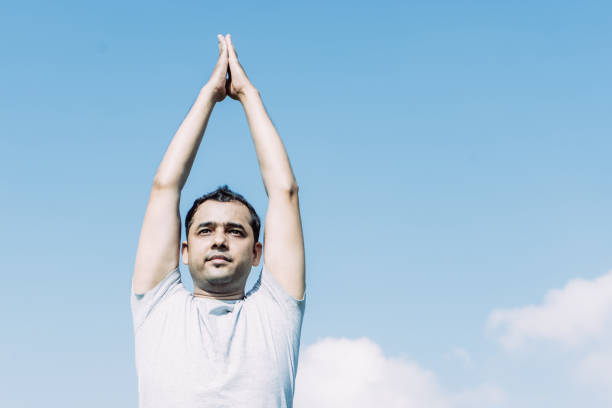 Indian man doing yoga and pressing hands together above head outdoors with blue sky in background. Outdoor yoga concept. Front view.