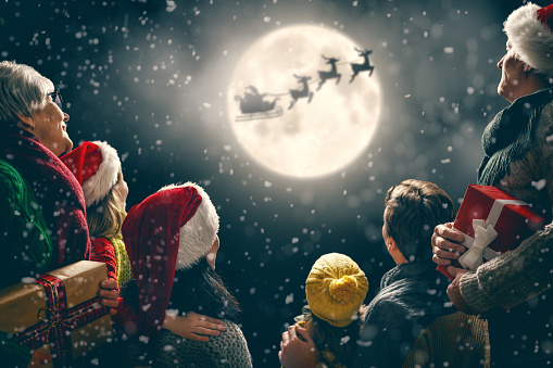 Merry Christmas and happy holidays! Cute little children with mom, dad, grandma and grandpa. Santa Claus flying in his sleigh against moon sky. Family enjoying the holiday on dark background.