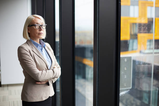istock Serious confident attractive mature lady in glasses standing at panoramic window in modern office and crossing arms on chest whole looking out window 1073477284
