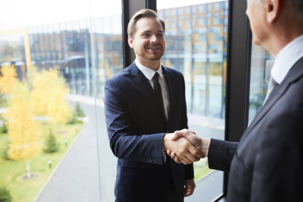istock Cheerful successful business partners in formal suits standing at panoramic window with view of city park and making handshake while congratulating each other with deal 1073477364