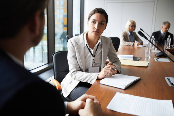 Serious attractive female journalist with badge hanging on neck sitting at table and making notes in diary while interviewing businessman at press conference Journalist interviewing businessman at press conference politician stock pictures, royalty-free photos & images