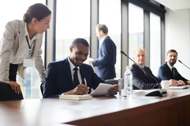 Photo of Content attractive young business lady with badge standing at black colleagues and giving advices him with papers while he analyzing information during conference break