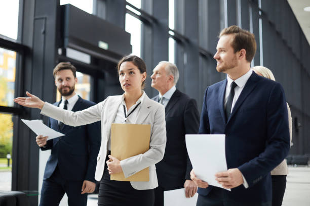 istock Serious confident female business guide in formal jacket holding big envelope and gesturing hand aside while showing direction to conference participants 1073477984