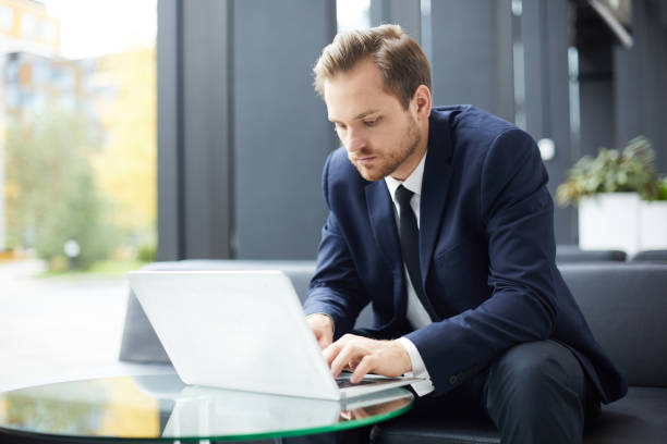 istock Serious busy handsome bearded entrepreneur in formal suit sitting on sofa and using laptop while composing email in hotel lobby with wifi 1073478032