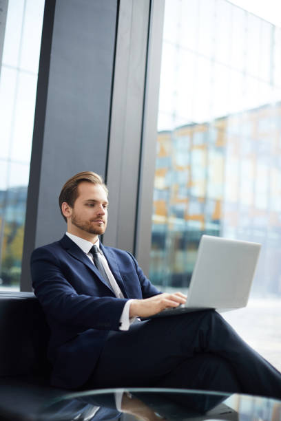 istock Content concentrated handsome bearded business manager sitting on sofa and preparing report on laptop while working in lobby 1073478162