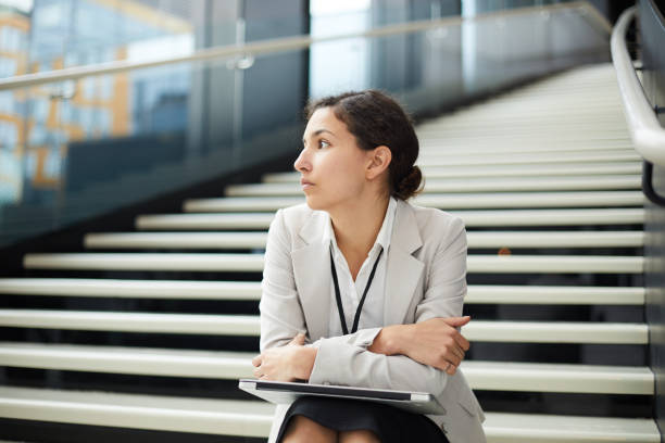 istock Sad pensive young business lady in jacket sitting on stairs and looking away while thinking about new projects and plans in hall 1073478342