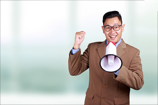 Young Asian businessman wearing suit using megaphone, shouting and smiling. Close up body portrait. Marketing promotion concept
