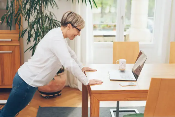 Fit mature businesswoman doing exercises in her office leaning forwards over the table reading her laptop with a smile