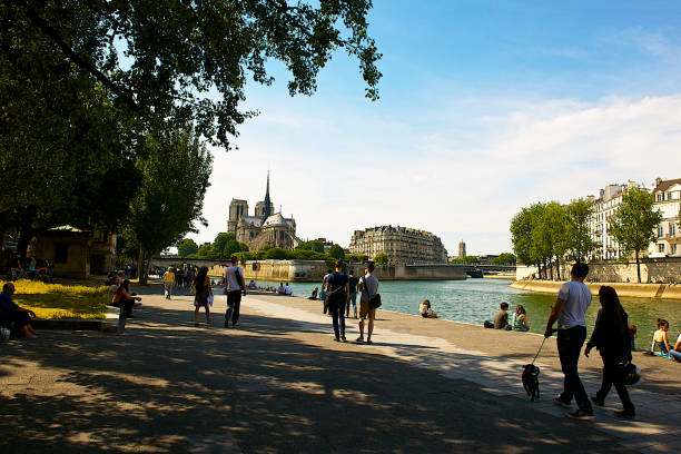 People walking along the left bank of the Seine river, on a summer afternoon, enjoying the view of the Notre Dame cathedral.