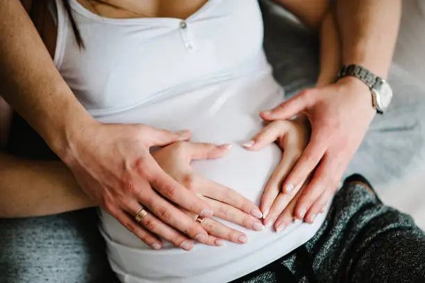 Photo of Cropped image of husband holding belly of his pregnant wife making symbol heart hands. Pregnant woman and loving handsome man hugging tummy at home. Loving Couple. Parenthood concept. Baby Shower.