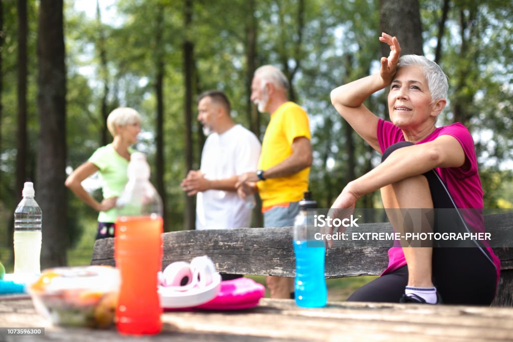 Active senior people taking a break after an exhausting exercise outdoors Group of active senior people taking a break after working hard exercising in a park. One woman in the front is sitting on a bench alone, resting with a leg up and wiping sweat off her forehead, while the rest are in a group behind her. 60-69 Years Stock Photo