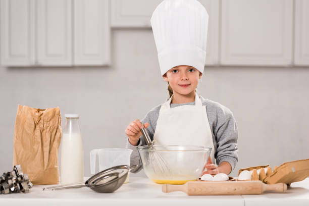 istock smiling child in chef hat whisking eggs in bowl at table in kitchen 1073605230