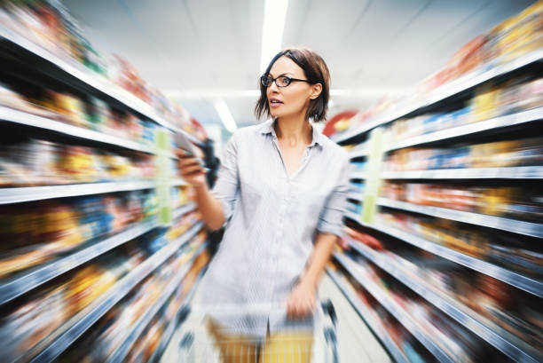 Grocery shopping. Attractive woman searching for food in a supermarket from a checklist on her phone. Motion blur background. checklist photos stock pictures, royalty-free photos & images