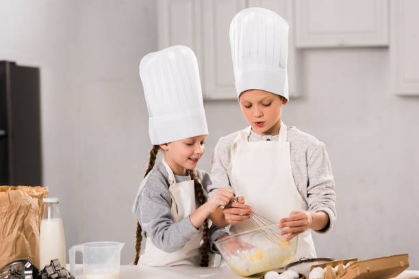 istock little kids in chef hats and aprons whisking eggs in bowl at table in kitchen 1073605246