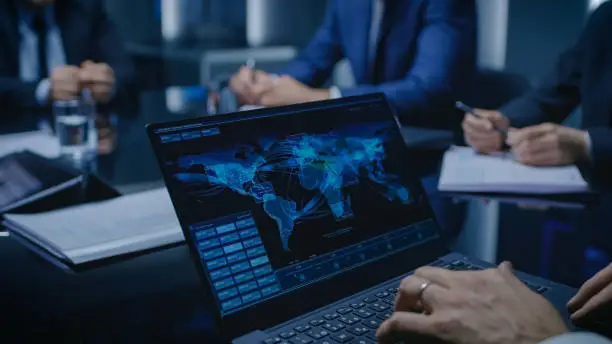 Photo of On the Table Laptop Showing World Data Flow: Team of Politicians, Corporate Business Leaders and Lawyers Sitting at the Negotiations Table in the Conference Room, Trying to Come to an Agreement.