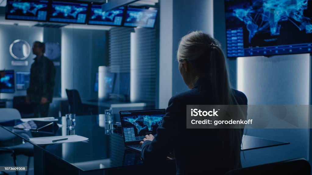Female Special Agent Works on a Laptop in the Background Special Agent in Charge Talks To a Military Man in the Monitoring Room. In the Background Busy System Control Center with Monitors Showing Data Flow. Military Stock Photo