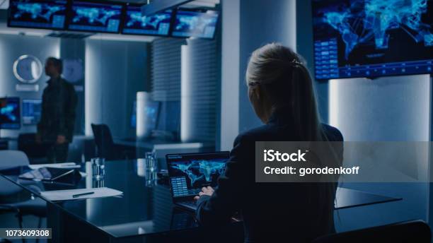 Female Special Agent Works On A Laptop In The Background Special Agent In Charge Talks To A Military Man In The Monitoring Room In The Background Busy System Control Center With Monitors Showing Data Flow Stock Photo - Download Image Now