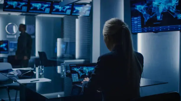Photo of Female Special Agent Works on a Laptop in the Background Special Agent in Charge Talks To a Military Man in the Monitoring Room. In the Background Busy System Control Center with Monitors Showing Data Flow.